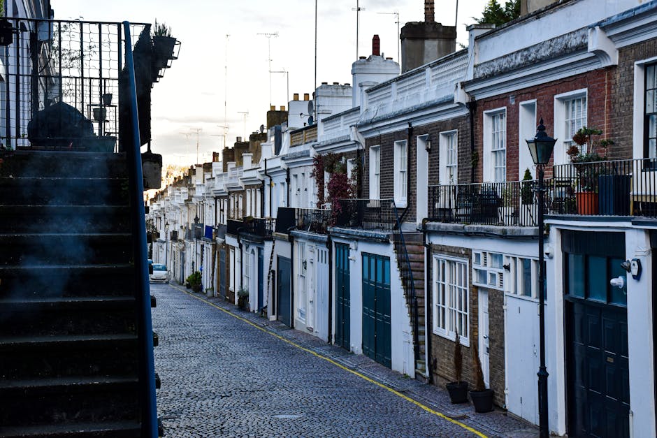 A row of traditional Georgian terraced houses in Holland Park, featuring brick facades with white wooden window frames and decorative cornices. Some houses have small front gardens with potted plants and outdoor lighting fixtures, while others have black iron balconies. The street is paved with cobblestones and is lined with black street lamps, with parked cars visible further down the road. On the left side, a black metal staircase with a mesh railing leads up to a house entrance, partially obscuring the view. The scene is captured during daylight hours with a soft, diffuse light, illustrating an urban residential area suitable for house removals and home relocation services. The image is associated with professional removals, packing, and furniture transport in Holland Park, as managed by Man with Van Holland Park.