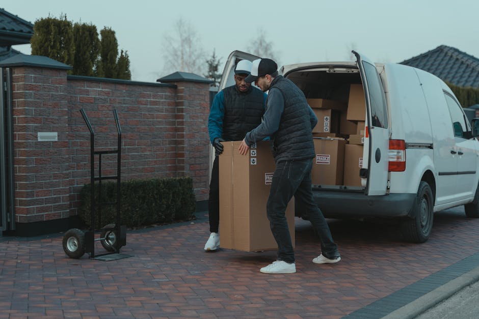 Two men are engaged in a furniture loading process outside a residential property, with one man lifting a large cardboard box onto a trolley and the other stabilising it. They are dressed in casual clothing suitable for manual work, including vests and gloves. The scene takes place on a paved driveway adjacent to a brick wall and a residential building with dark tiled roofing. Behind them, a white van with its rear doors open reveals several packed cardboard boxes inside, ready for transport as part of a home relocation or moving service. The men are positioned next to the van, which is parked on the driveway, and additional packing materials such as cardboard boxes and protective blankets are visible. This image illustrates a professional furniture transport and packing and moving process, highlighting the loading stage of a house removal conducted by Man with Van Holland Park.
