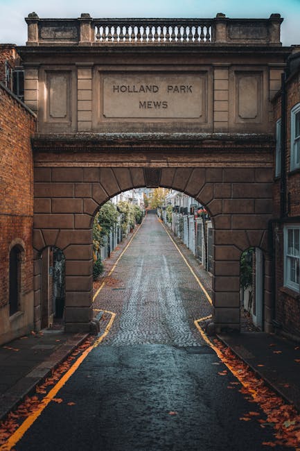 A narrow cobbled street seen through an arched stone gateway inscribed with 'HOLLAND PARK MEWS', flanked by brick and stone buildings. The street extends into the distance, lined with residential fences, trees, and parked cars, with fallen leaves on the pavement indicating an autumn setting. Part of a house removal or furniture transport process is inferred from the context of the scene, although no moving equipment or personnel are visible in the image. The scene is captured during daytime with natural lighting illuminating the historic gateway and the street beyond, consistent with a typical London residential area near Holland Park. This environment is representative of a location where professional house removals, such as those by Man with Van Holland Park, might operate, involving meticulous packing and transportation of household items through the quaint mews.