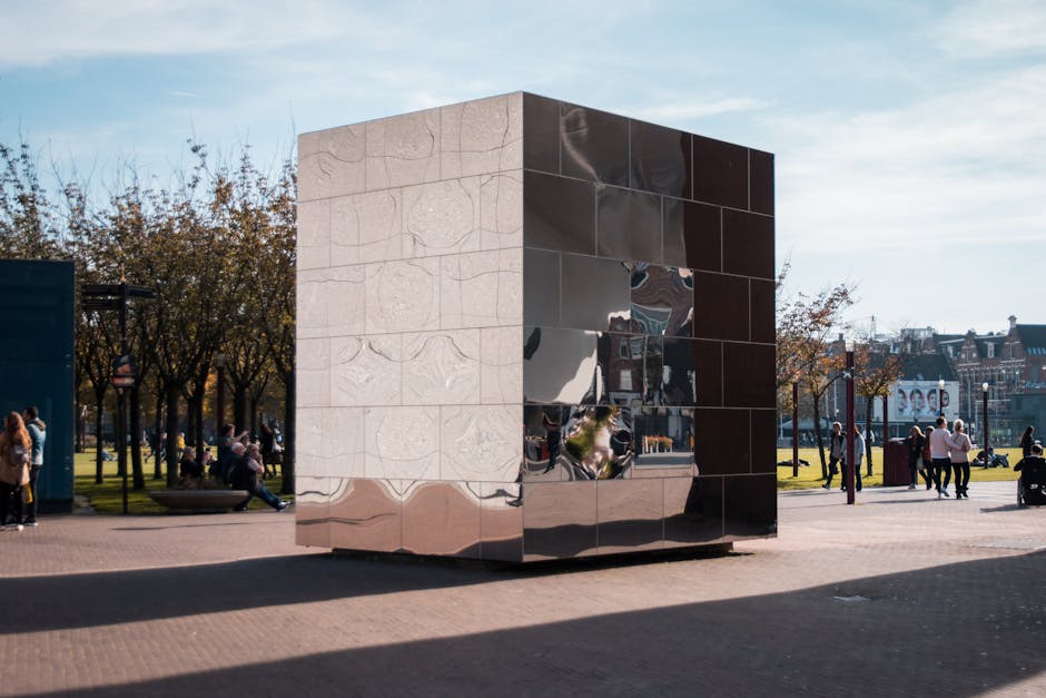 A large reflective cube sculpture composed of a combination of shiny metallic and light-colored stone or concrete panels is situated outdoors on a paved area. The sculpture's surface features smooth, polished facets and visible seams between the panels. It is positioned on a flat, level surface with a shadow cast underneath. In the background, there is a park with several leafless trees, some people sitting on benches, and groups walking or standing in small clusters. A clear sky with some light clouds is visible, suggesting daytime with ample natural light. The scene appears to be in an urban public space, possibly a city park or plaza, with buildings and billboards visible in the distance. This setting includes elements typical of an active pedestrian area, which Man with Van Holland Park may utilise for house removals or furniture transport in busy environments.