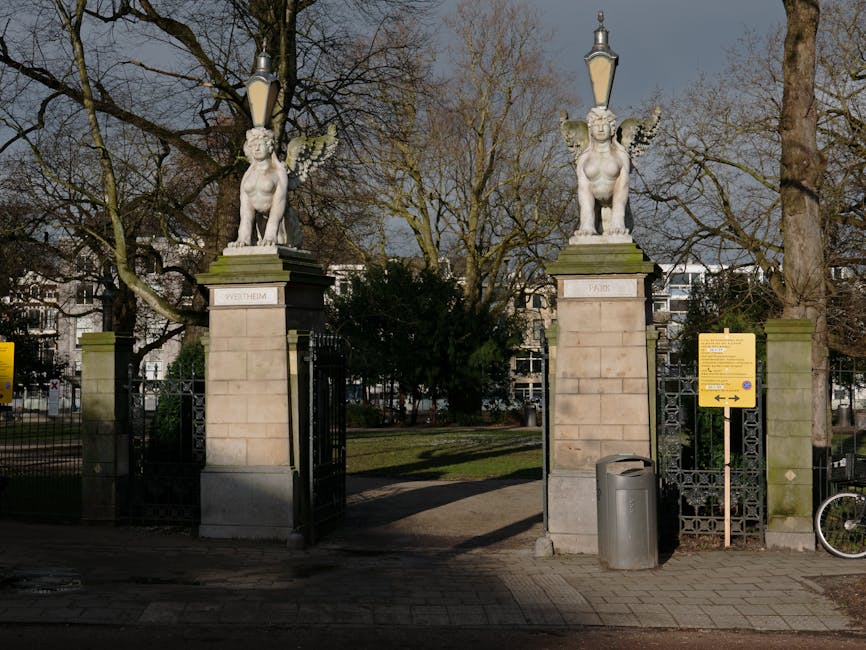 Image of a park entrance with two stone pillars, each topped with a white stone statue of a winged figure resembling a mythological creature, with a city street in the background. The left pillar has a plaque with the word 'WETHEIM', and the right with 'PARK'. Black wrought iron gates are open, revealing trees with bare branches and some green shrubbery inside the park. To the right of the entrance, there is a gray metal trash bin and a partially visible bicycle leaning against a metal post. An informational yellow sign with black text is mounted on a post next to the gate. The pavement outside the entrance is made of gray bricks, and the scene appears to be taken during daylight with an overcast sky. The context suggests a location that could be involved in home relocation or moving logistics, which is relevant to services like [COMPANY_NAME] as they assist with house removals in the area.