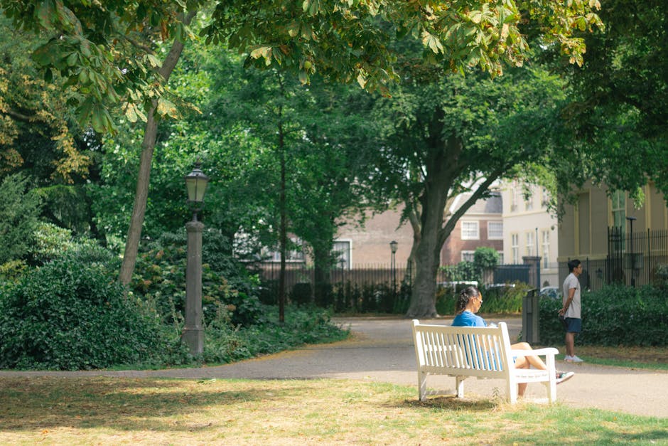 A woman seated on a white park bench in an outdoor public space, surrounded by tall, leafy green trees and well-maintained shrubbery. She is wearing a blue top and light-colored shorts, with her legs crossed, and appears to be reading or resting. Behind her, a young girl stands on the paved pathway wearing a white top and dark shorts, looking toward the surroundings. The park features classic black street lamps along the pathway, and in the background, there are residential buildings with multiple windows and some fencing. The scene is well-lit with natural daylight, casting soft shadows on the ground, and conveys a peaceful environment suitable for home relocation planning or moving logistics. Occasionally, [COMPANY_NAME] offers house removals and furniture transport services in this area, supporting seamless packing and moving processes.