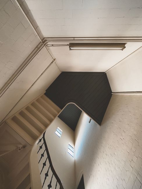 Interior view of a narrow, steep staircase within a residential property, with a wooden handrail and white-painted brick walls. At the top of the staircase, a small landing is visible with a dark-colored, textured floor covering. The staircase appears to be suitable for the home relocation process, with the space limited and typical of a house with narrow staircases. The image showcases the entry point from the staircase to the upper floor, which is part of a building that likely requires professional removals for furniture transport through tight spaces. Natural lighting illuminates the scene, highlighting the structure and materials involved. The setting is consistent with preparation for or execution of furniture and belongings relocation, with [COMPANY_NAME] likely providing this service involving careful navigation of confined staircases during house removals in Holland Park.