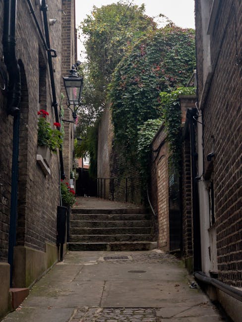 Image of a narrow, outdoor stairway between two brick buildings in an urban setting, leading upward to a small landing surrounded by greenery, including large bushes and climbing plants. The stairway features concrete steps with metal handrails on both sides, and the ground in the foreground is paved with stone slabs. The scene is lit with natural daylight, highlighting the aged brickwork and foliage. This setting illustrates a typical residential alleyway that might be part of a home relocation involving narrow stair removals, a service offered by Man with Van Holland Park, with packing and furniture transport carefully managed through tight spaces.
